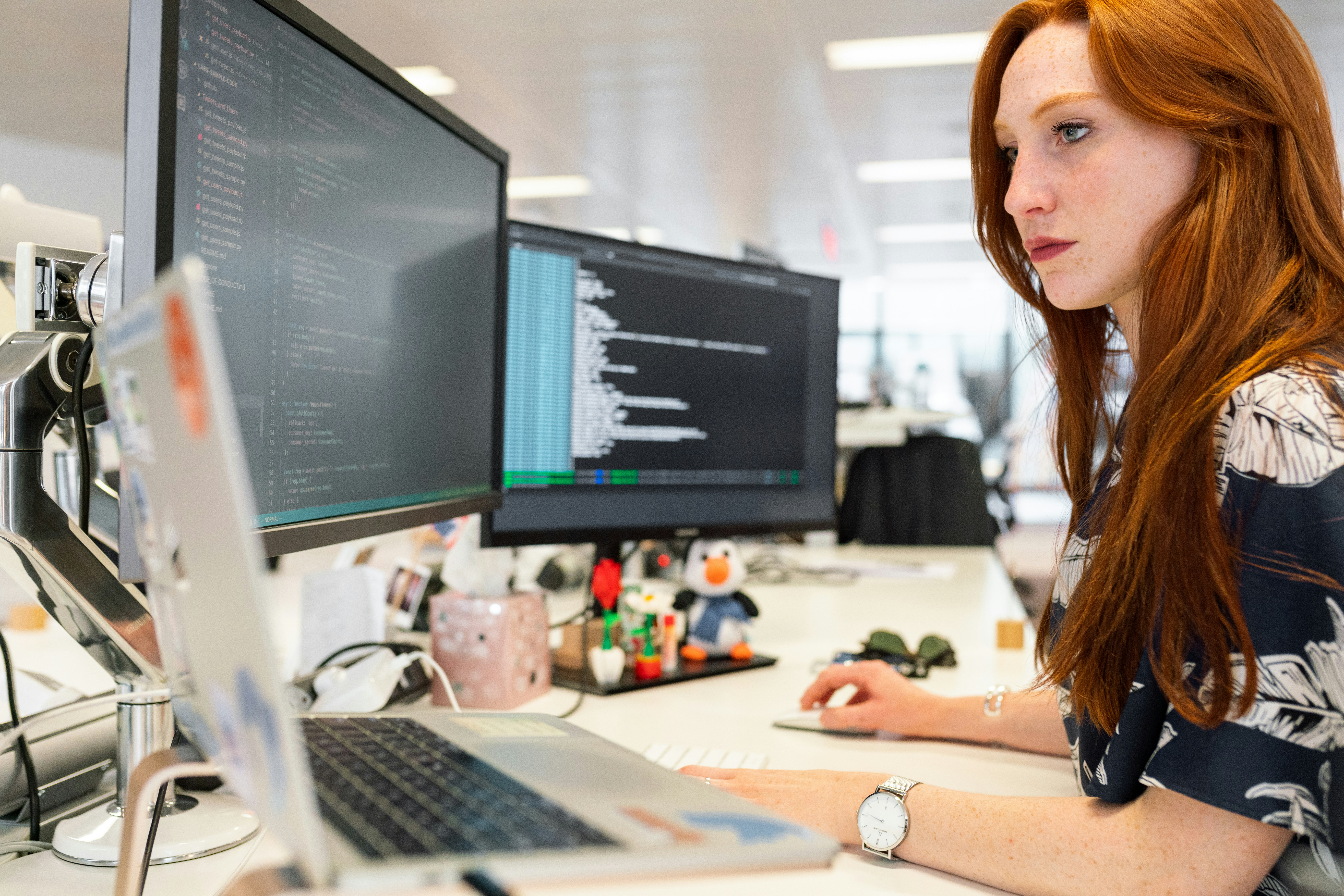 Woman with red hair seated at computer with dual monitors and her head turned left.